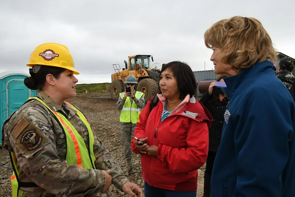 Three people, one in fatigues and hard hat, standing in front of bulldozer discussing relocation plans in Newtok, Alaska.